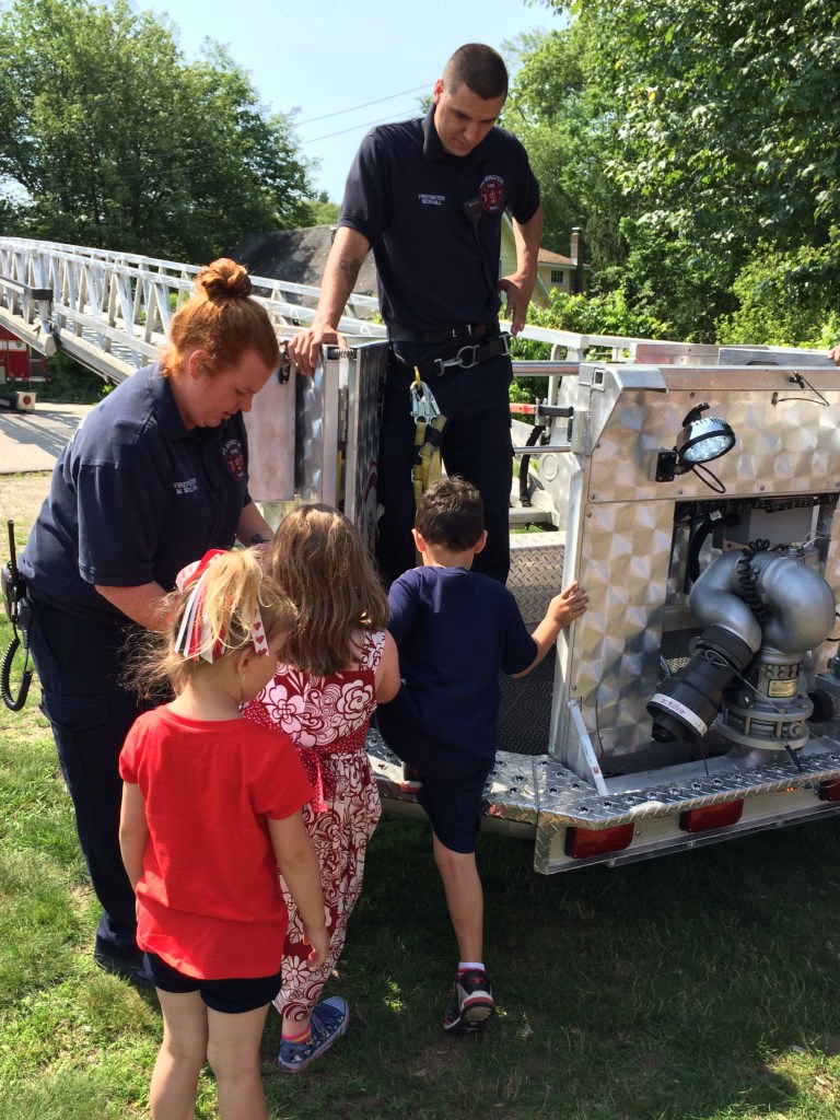 Children from the Tiny Tots/Kids Club program explore the bucket from the  Engine 1 Ladder Truck with the assistance of Firefighter Sullivan and Firefighter Newhall. 